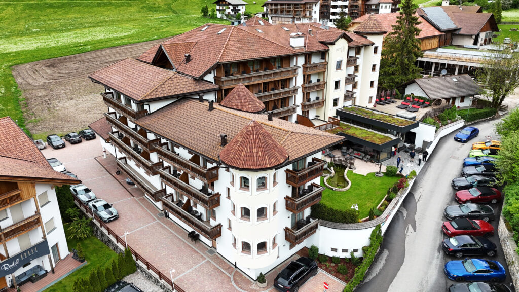 Luftaufnahme eines Hotelgebäudes, des Almhof Hotel Call, mit angrenzender Straße, an der Mustangs geparkt sind.