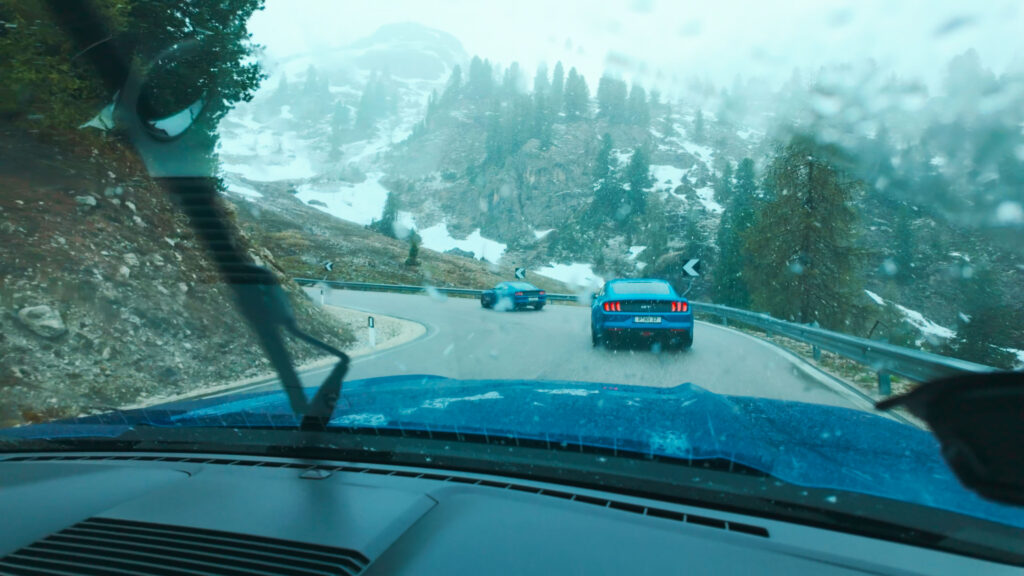 Blauer Mustang am Rand einer Straße mit schneebedeckten Bergen im Hintergrund.