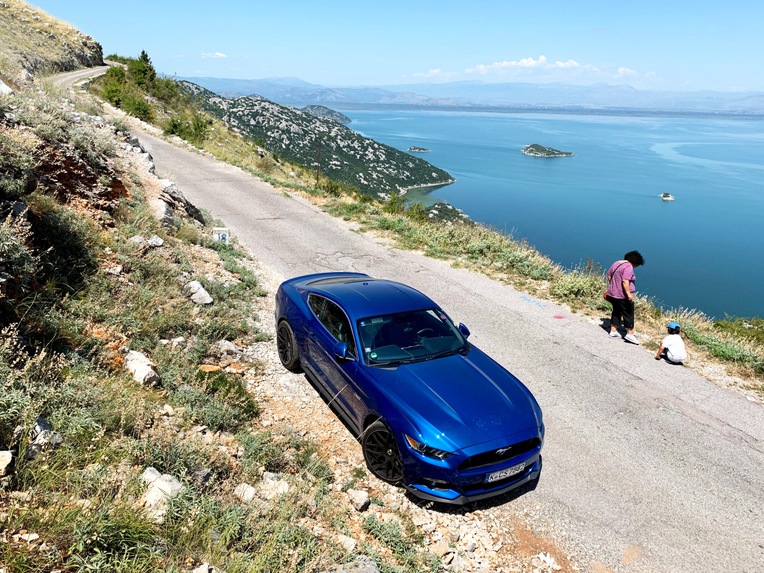 Blauer Mustang parkt auf einer schmalen Küstenstraße in Montenegro mit Blick auf das Meer.
