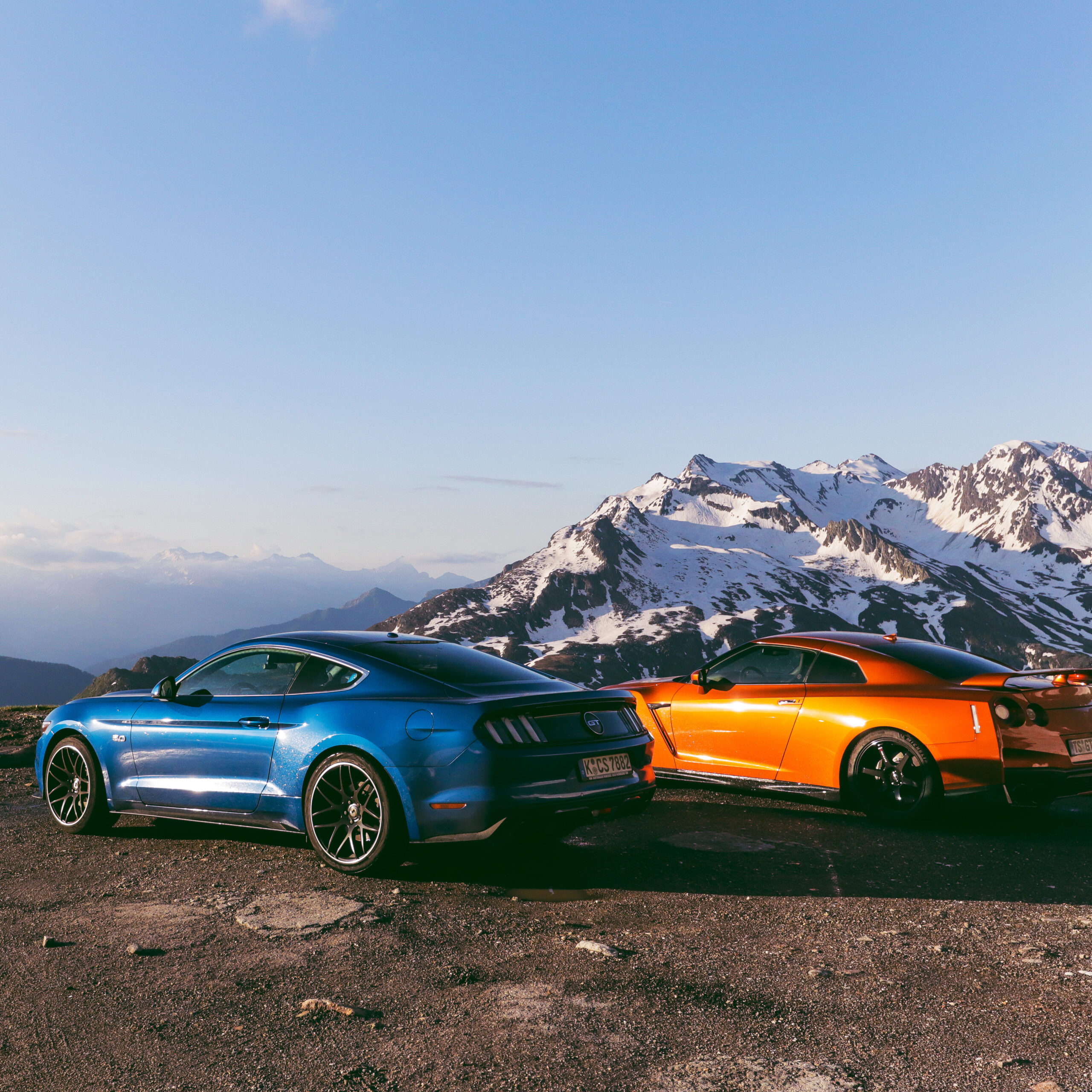 Blauer Mustang GT und orangefarbener Nissan GT-R am Timmelsjoch vor verschneiten Bergen im Sonnenuntergang.