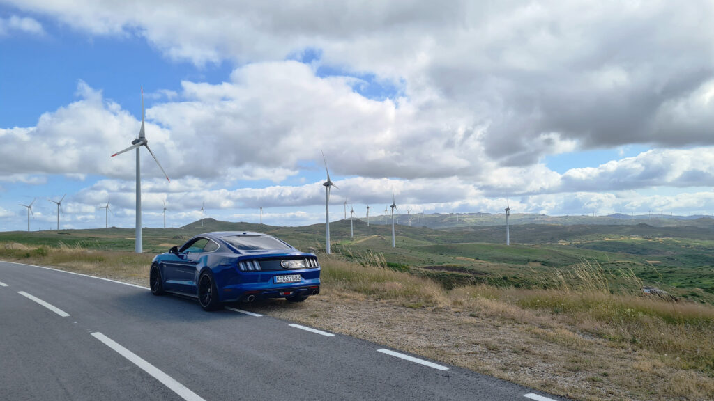 Blauer Mustang steht am Straßenrand in Portugal mit Blick auf Windräder und Hügel.